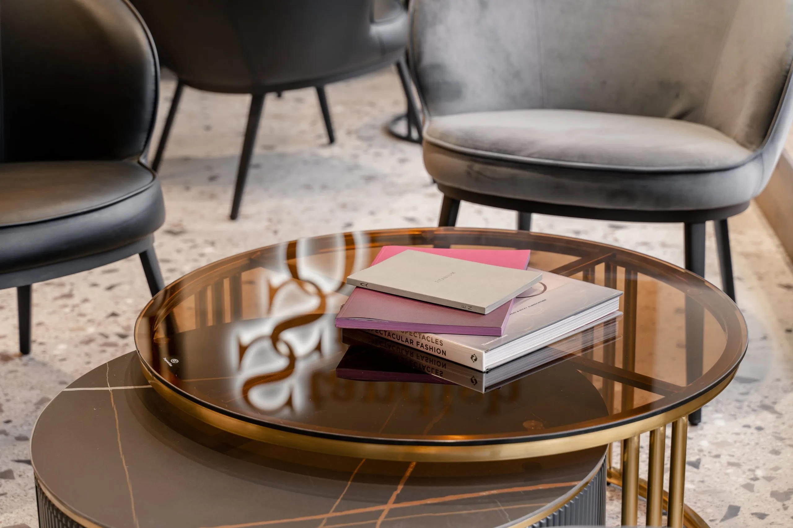 Fashion books on a smoked glass table between grey velvet chairs
